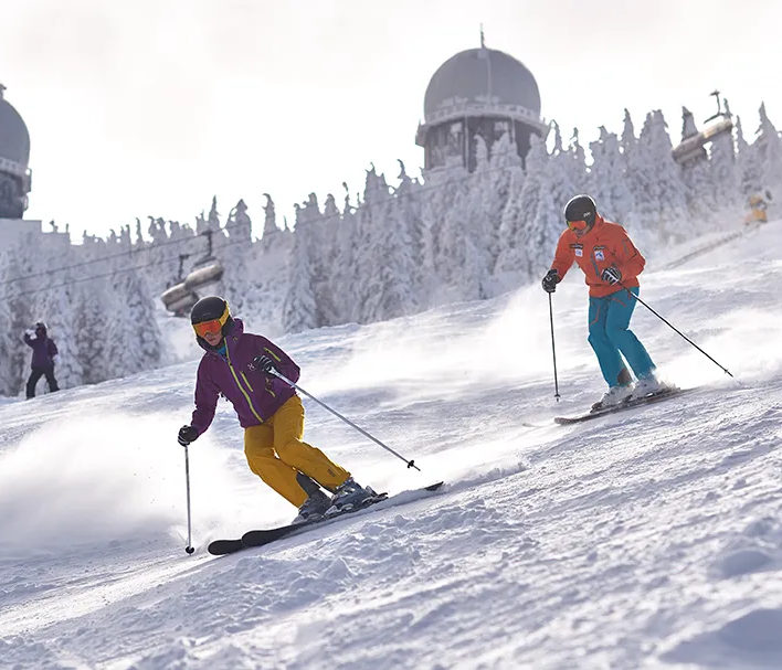 Zwei Skifahrer gleiten mit Schwung die verschneite Piste am Großen Arber hinunter. Im Hintergrund die Radarkuppeln.