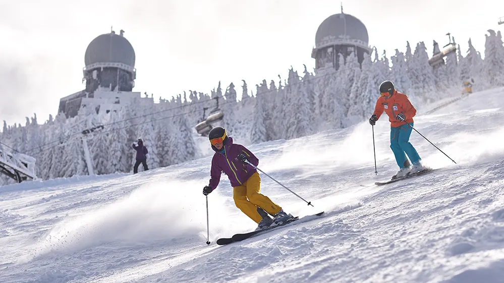 Zwei Skifahrer gleiten mit Schwung die verschneite Piste am Großen Arber hinunter. Im Hintergrund die Radarkuppeln.