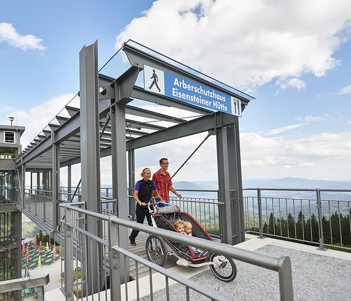 Auf der hohen modernen Aussichtsplattform mit Aufzug spaziert ein Paar mit einem Kinderwagen, während sich dünne Wolken über den Bergen formieren.