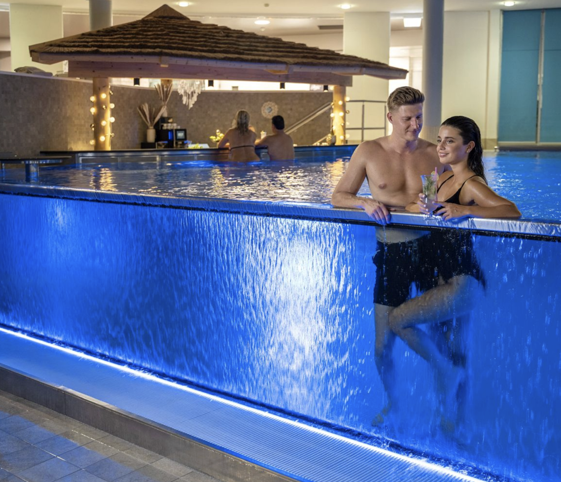 Im Hintergrund befindet sich eine mit Schilf bedeckte Bar, während das Paar in einem blauen, beleuchteten Pool entspannt.