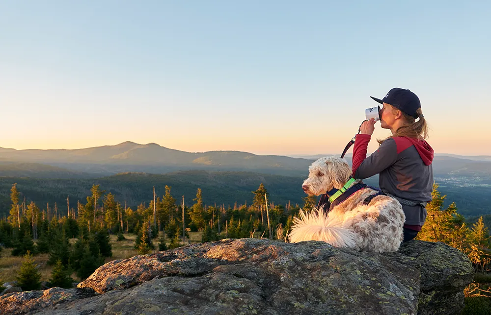 Eine Frau trinkt genüsslich aus ihrem Becher, auf einem Bergvorsprung sitzend, während ihr Hund neben ihr liegt und mit die Auszeit genießt.