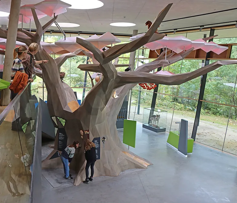 Ein begehbarer, stilisierter Baum steht im hellen Ausstellungsraum, während Besucher ihn neugierig erforschen.