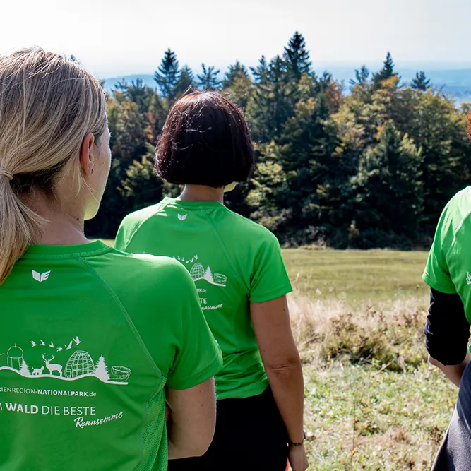 Drei Frauen in grellgrünen FNBW-T-Shirts stehen erhaben auf einer Wiese am Almberg und blicken auf die sich ihnen eröffnende weite Aussicht ins Vorland.