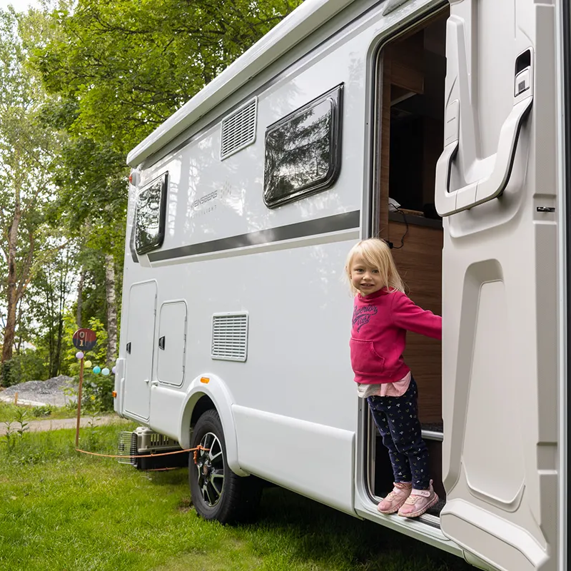Das blonde Mädchen in einem pinken Pullover lächelt, während sie aus der offenen Tür eines modernen Wohnmobils blickt.