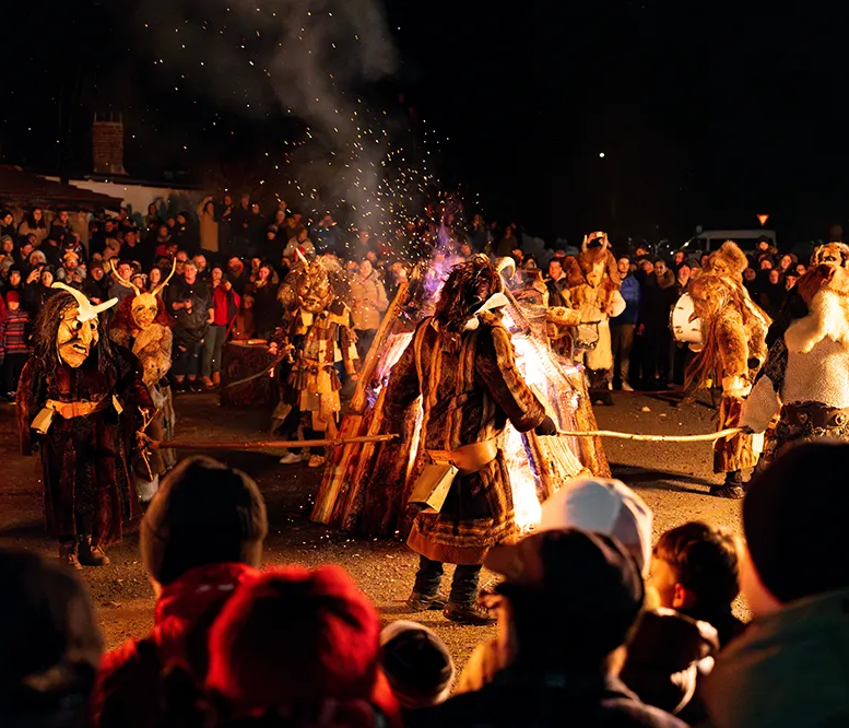 Die Szene zeigt maskierte Lousnachtsgeister, die um ein loderndes Feuer herumtanzen, während eine Menschenmenge zuschaut.