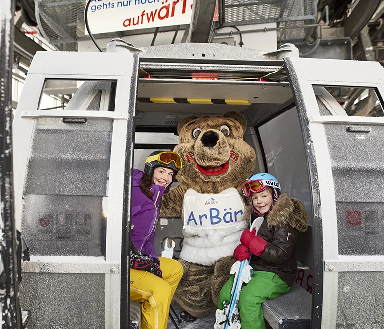 In einer Gondel sitzen zwei Skifahrer-Mädels und ein Bärenmaskottchen, bereit um auf den Berg zu starten für ein Abenteuer im Schnee.