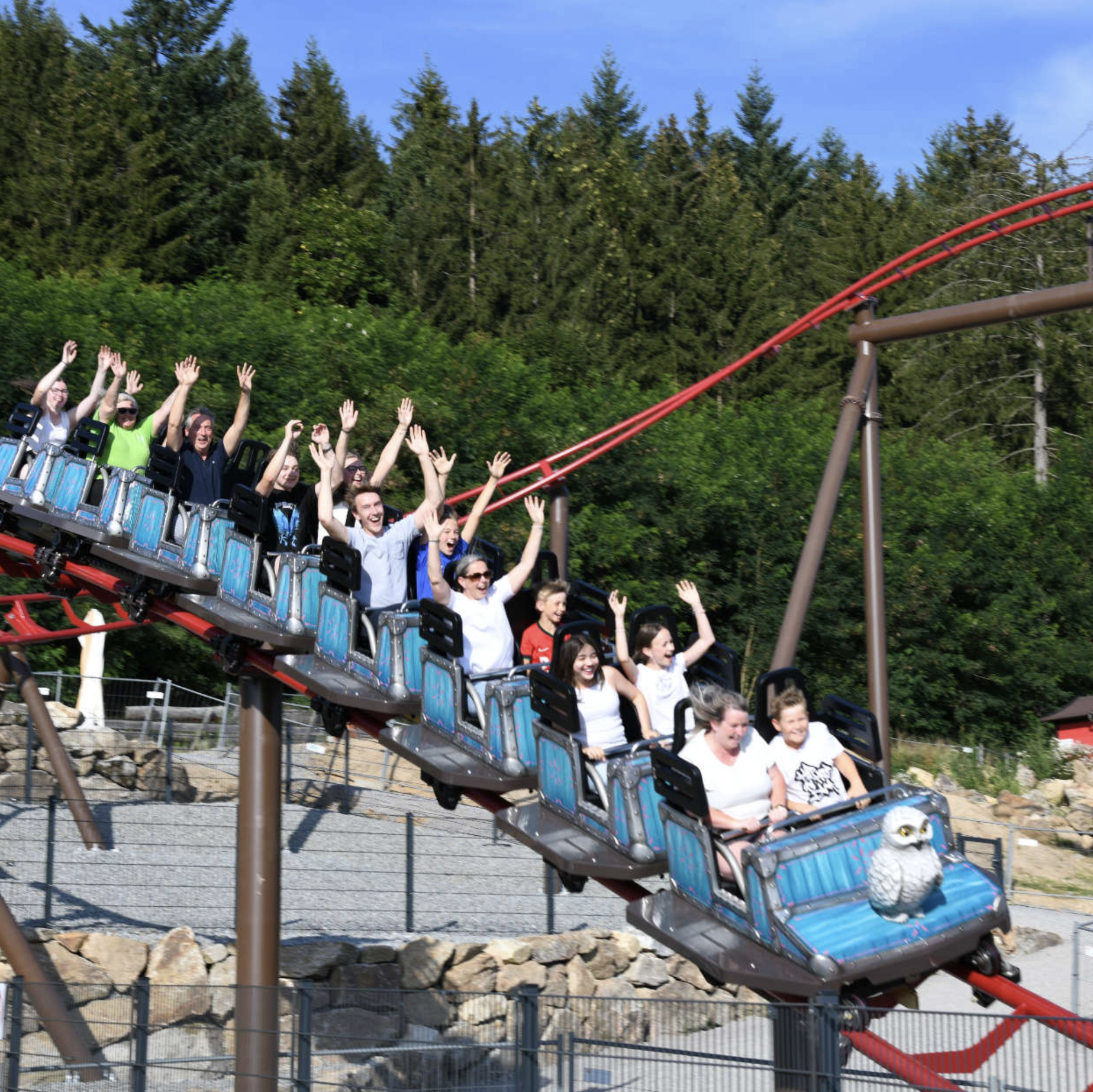 In einem Freizeitpark genießen viele Menschen die Fahrt auf einer Achterbahn, umgeben von Bäumen und einer sonnigen Atmosphäre.
