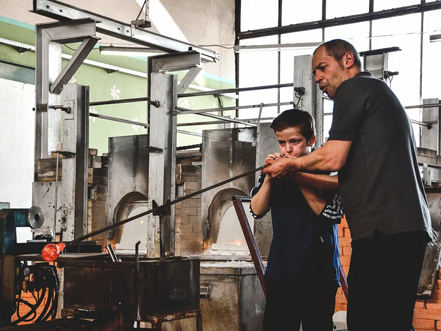 Die Szene zeigt einen Mann, der einem Kind beim Glasblasen hilft, während sie in einer typischen Glashütte stehen.