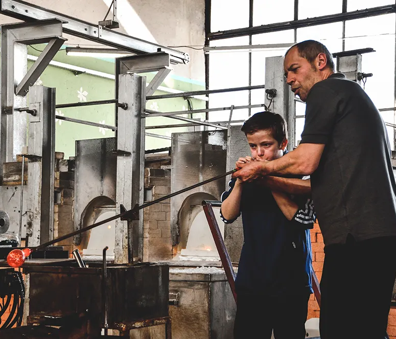 Die Szene zeigt einen Mann, der einem Kind beim Glasblasen hilft, während sie in einer typischen Glashütte stehen.