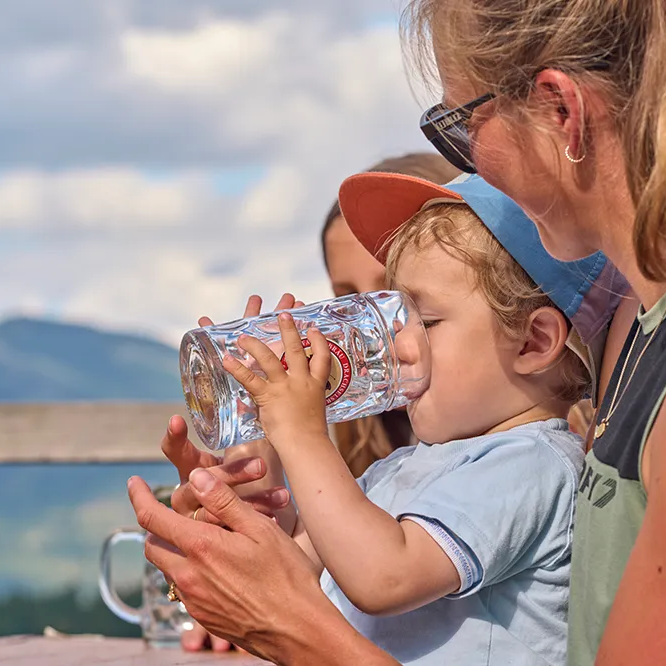 Ein kleiner Junge stillt seinem Durst und trinkt aus einem großen Glas, während er von einer Frau mit langen Haaren umgeben ist, die ihn liebevoll unterstützt.