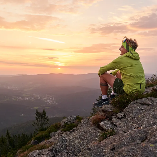 Die goldenen Sonnenstrahlen tauchen die Berge in warmes Licht, während ein Mann entspannt auf einem Felsen sitzt mit Blick in die Ferne.