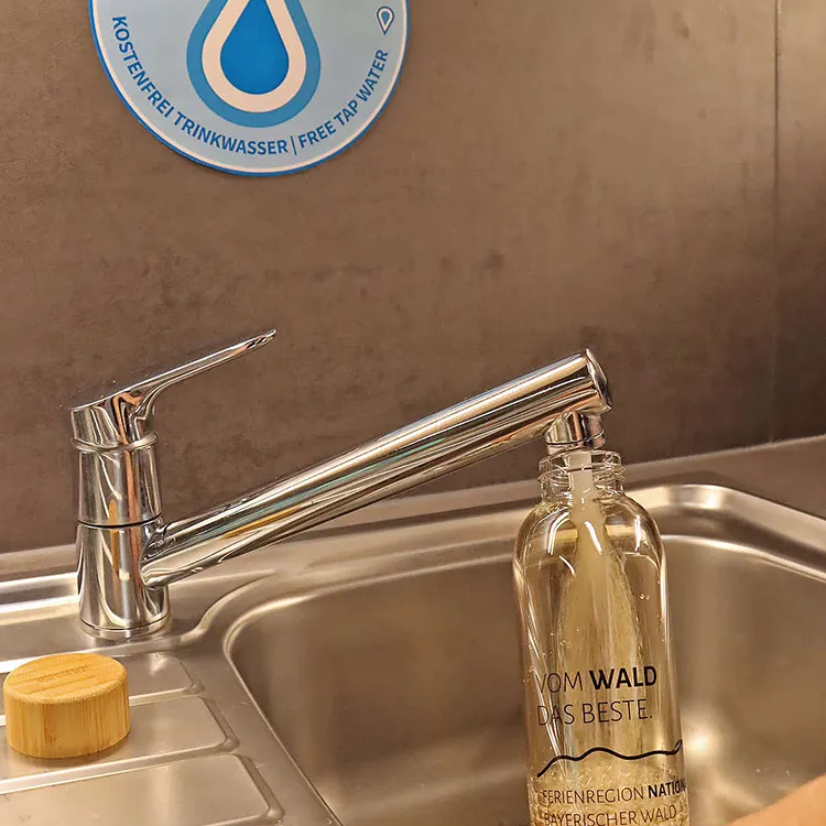 Wasserauffüllstation "Refill" für umweltfreundliche Trinkgewohnheiten Der Wasserhahn sprudelt frisches Waldwasser in die nachhaltige Glasflasche, während ein Schild auf die kostenlose Trinkstation hinweist.