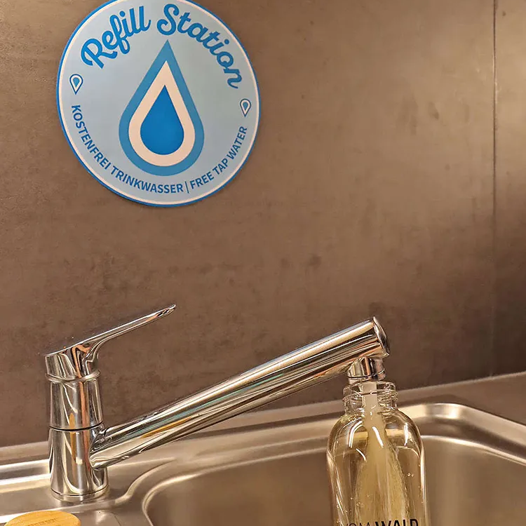 Der Wasserhahn sprudelt frisches Waldwasser in die nachhaltige Glasflasche, während ein Schild auf die kostenlose Trinkstation hinweist.