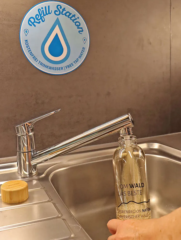 Der Wasserhahn sprudelt frisches Waldwasser in die nachhaltige Glasflasche, während ein Schild auf die kostenlose Trinkstation hinweist.