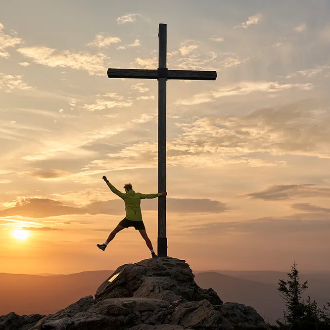 Vor einem imposante Sonnenuntergang steht eine Mann in Outdoorkleidung am Gipfelkreuz auf einem Felsen und hebt beschwingt einen Arm und ein Bein zur Seite.
