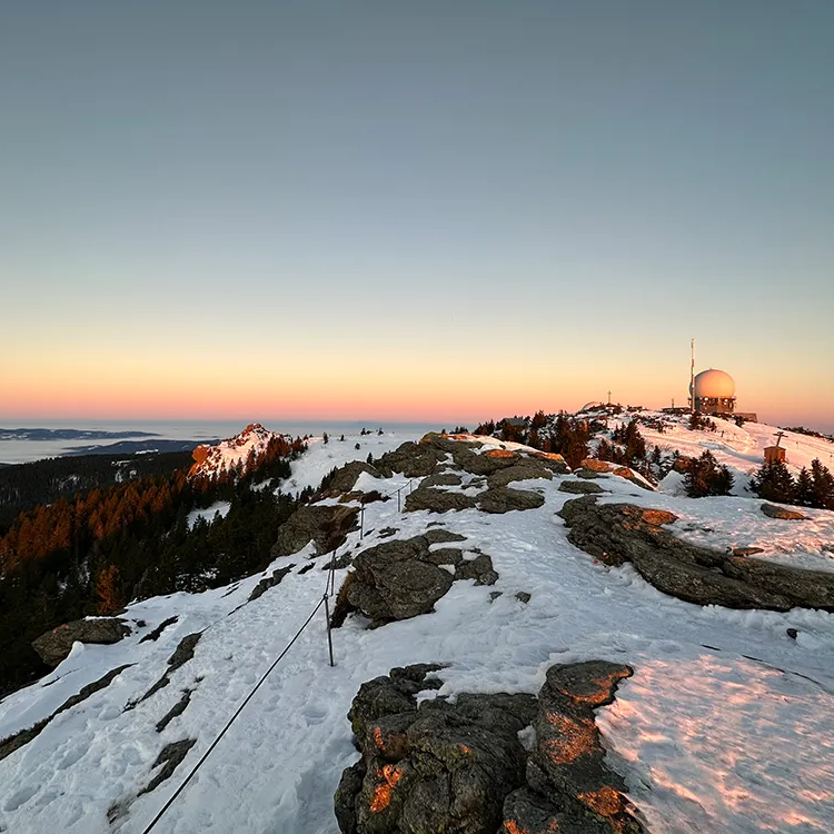 Der Sonnenuntergang färbt den Himmel über dem schneebedeckten Gipfel, während der Radarturm im Hintergrund sichtbar ist.