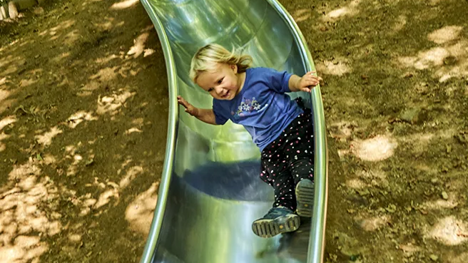 Ein kleines blondes Kind mit einem breiten Lächeln rutscht auf einer metallischen Rutsche, die auf einem Erdhügel angebracht ist.