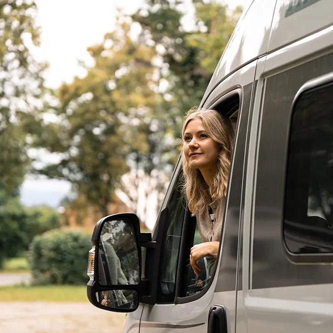 Eine blonde Frau genießt die Aussicht aus dem Fenster eines grauen Wohnmobils, während sie in der Natur parkt.