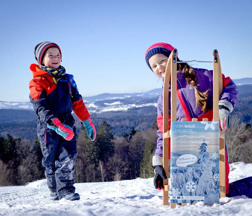 Zwei Kinder stehen fröhlich im Schnee, eines hält einen Schlitten mit einem Werbeschild für die Ferienregion Nationalpark Bayerischer Wald in den Händen