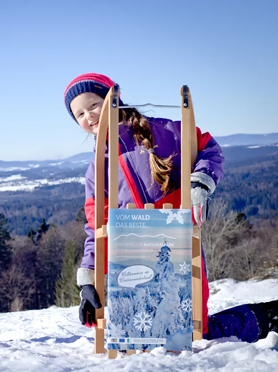 Zwei Kinder stehen fröhlich im Schnee, eines hält einen Schlitten mit einem Werbeschild für die Ferienregion Nationalpark Bayerischer Wald in den Händen