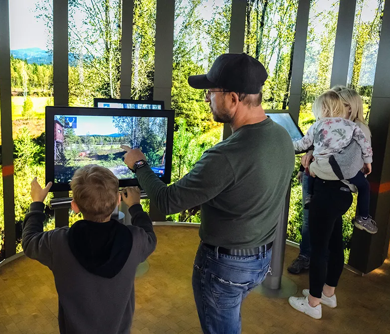 Ein Vater zeigt seinem Sohn auf einem Bildschirm eine Landschaft, während seine Frau mit der Tochter im Hintergrund steht.