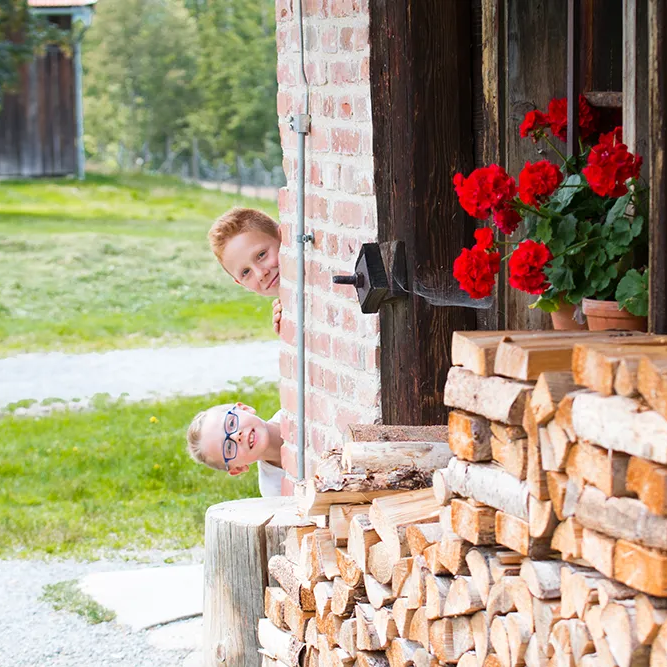 Zwei Kinder schauen spitzbübisch um die Ecke eines Holzhauses, vor dem aufgerichtetes Holz und leuchtend rote Geranien zu sehen sind.