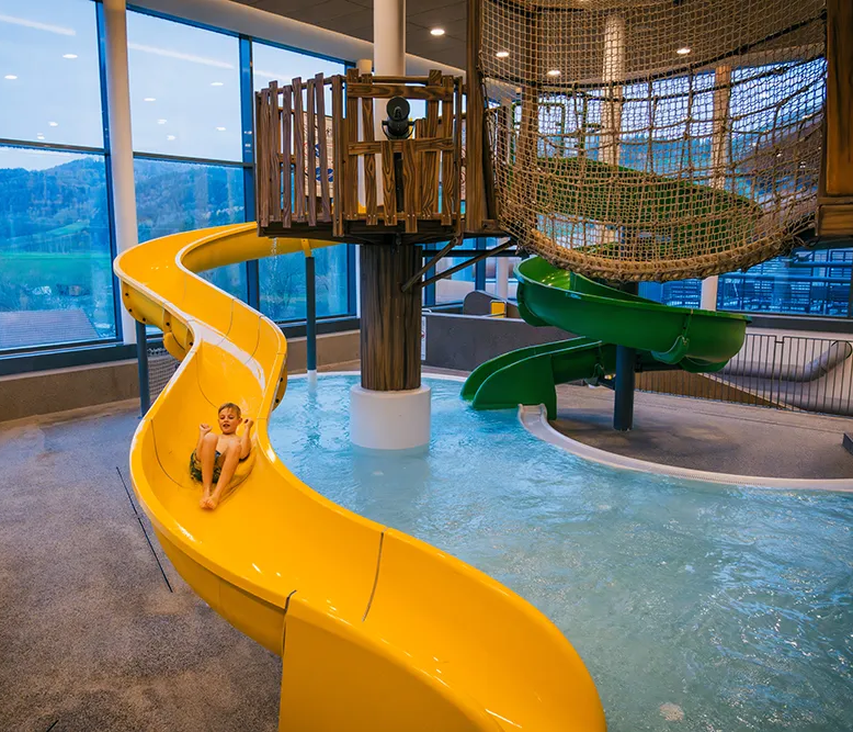 Im Hallenbad des Schreinerhofes, rutscht ein Kind mit Begeisterung die gelbe Rutsche hinunter, während die Landschaft draußen sichtbar ist.