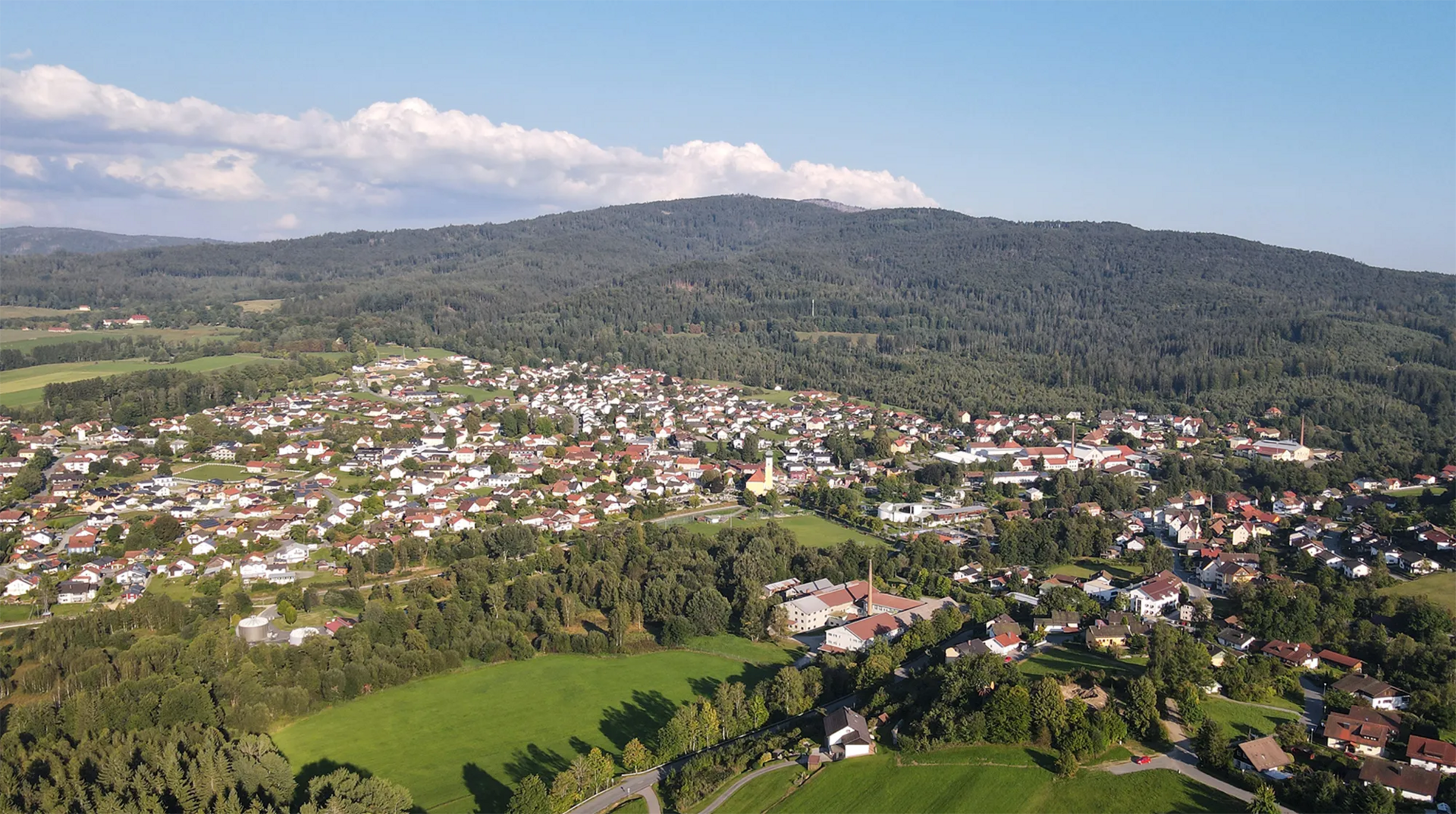 Luftansicht des Gläsernen Herzens, des charmanten Ortes Frauenau, das sich harmonisch in die Berg-Wiesen-Welt einfügt, mit einem klaren blauen Himmel und sanften Wolken.