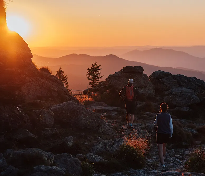 Der Mann mit Rucksack und die Frau, nur noch in schattenhaften Silhouetten, wandern den felsigen Weg hinauf, während die Sonne eine goldene Abendstunde schafft.
