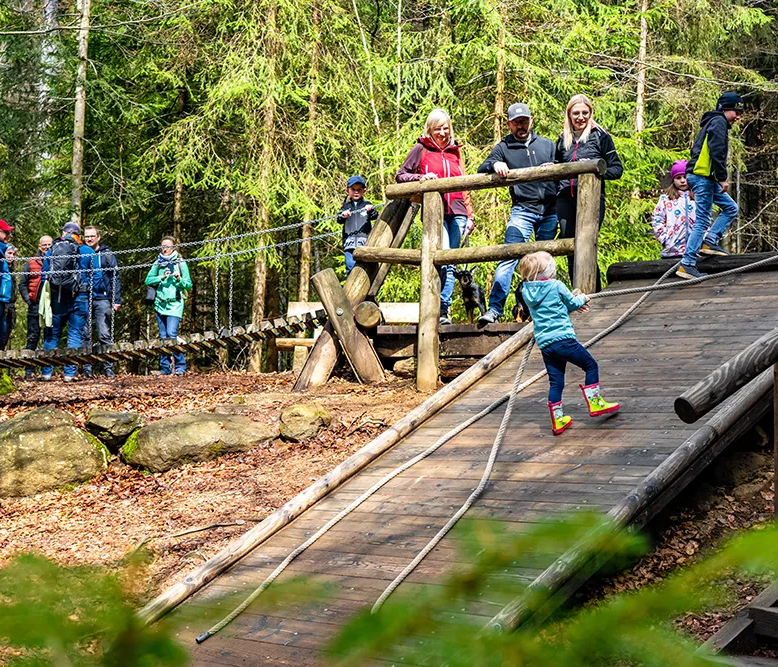 Ein kleines Kind in bunten Gummistiefeln klettert fröhlich die Holzrampe am Spielplatz im Wald hinauf, während Erwachsene zuschauen.