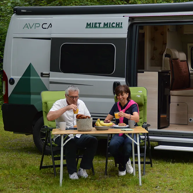Das Paar sitzt in Campingstühlen vor ihrem Wohnmobil und genießt ein Picknick mit frischen Lebensmitteln und Getränken am Tisch.