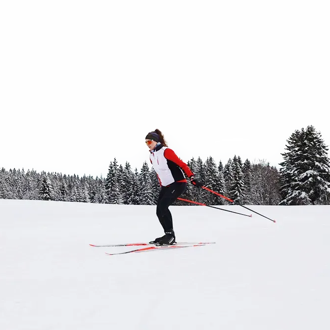 Eine sportliche Frau in Skikleidung bewegt sich auf Langlaufskiern elegant über den Schnee, umgeben von einer ruhigen, verschneiten Waldlandschaft.