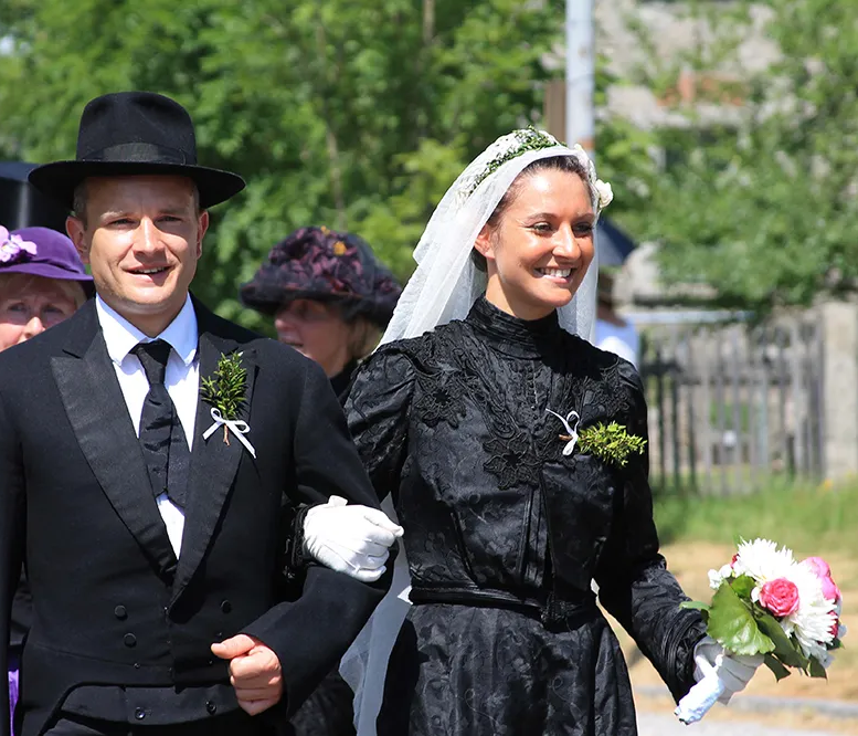 Die Braut trägt am Heimatfest Mauth ein elegantes historisches schwarzes Kleid und einen Schleier, während sie mit ihrem Bräutigam lächelt und Blumen hält.