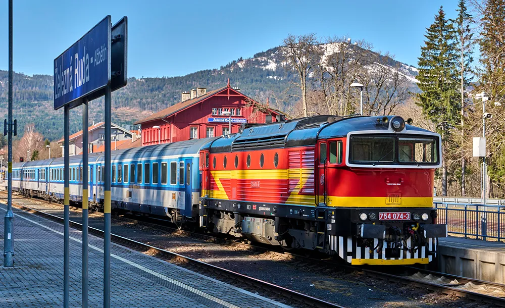 An einem sonnigen steht der farbenfrohe Zug der tschechischen Bahn am Grenzbahnhof, dahinter der markante Große Arber im Blickfeld..