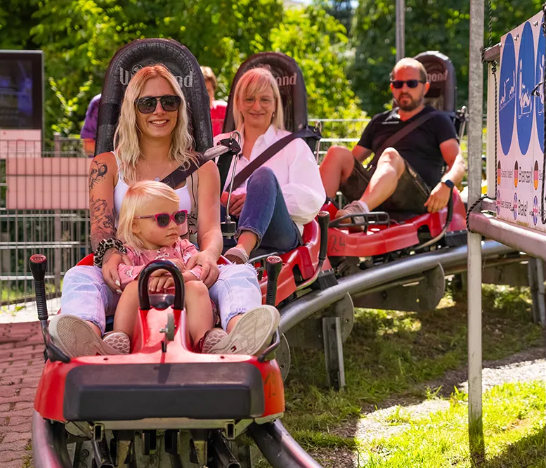 Spaßige Rodelpartie mit Kind an einem sonnigen Tag im Bärenbob Die Familie lächelt und entspannt sich in ihren Rodeln, während sie auf den Start der Abfahrt des Bärenbob in Grafenau wartet.