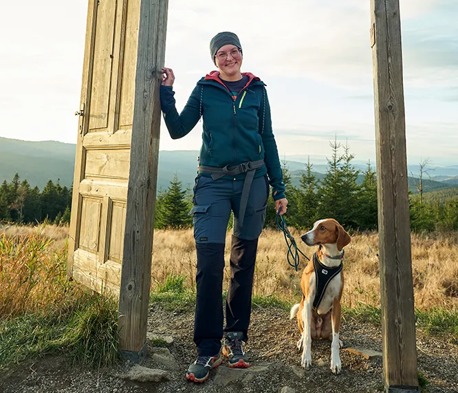 Eine Frau in Wanderkleidung steht lächelnd in einem Holzrahmen mit der Aufschrift 'WeLoveSumava', während ihr Hund neben ihr sitzt.