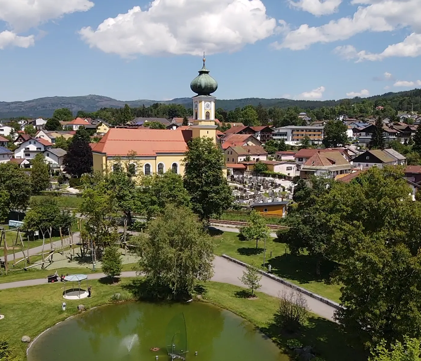 Die Rokokokirche mit der grünen Kuppel erhebt sich über Frauenau, während sich in den Gläsernen Gärten der Weiher im Vordergrund zeigt.