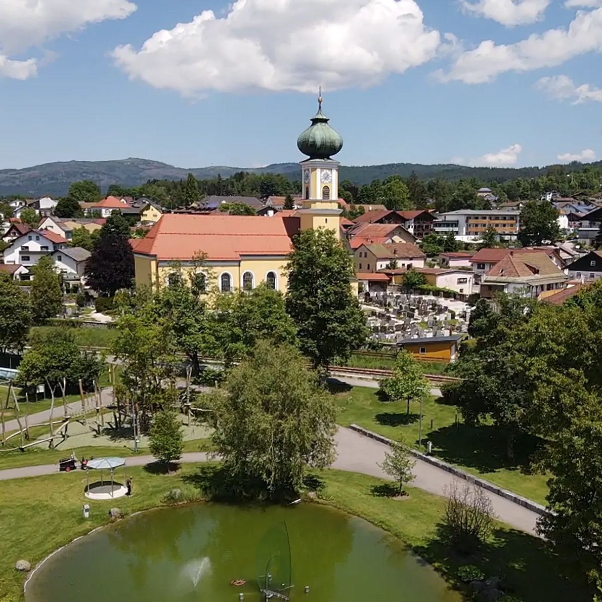 Malerisches Zentrum von Frauenau mit Rokokokirche und grünen Parkanlagen Die Rokokokirche mit der grünen Kuppel erhebt sich über Frauenau, während sich in den Gläsernen Gärten der Weiher im Vordergrund zeigt.