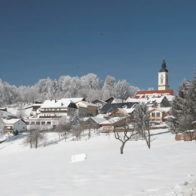 Winterzauber: Kirchdorf im Wald in der verschneiten Natur Kirchdorf im Wald im Winter, mit schneebedeckten Dächern und einer markanten Kirchturmspitze, die in den blauen Himmel ragt.