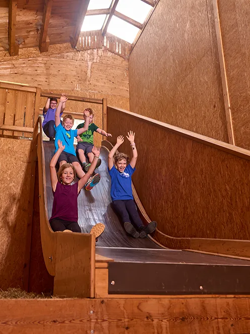 Fünf Kinder rutschen in einer Spielscheune fröhlich eine große Holzrutsche hinunter, während sie die Hände in die Luft werfen und sich freuen.