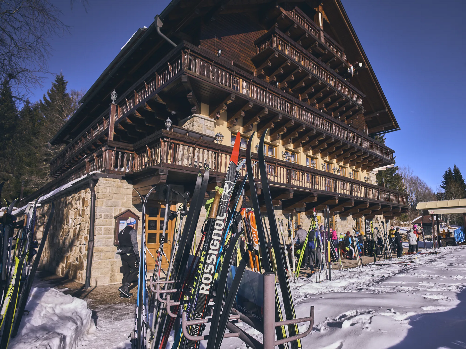 Das Bild zeigt ein rustikales Skihütte mit Holzverkleidung, umgeben von Schnee und Langläufern, die einen Langlauftag an der Grenze zum Sumava genießen.