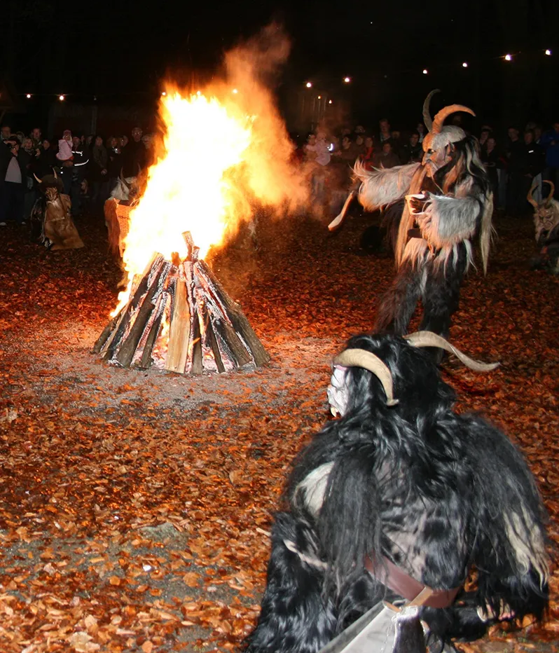 Begeisterte Besucher versammeln sich um ein großes Feuer, während mystische Rauhnachtsfiguren mit Hörnern im Vordergrund stehen.