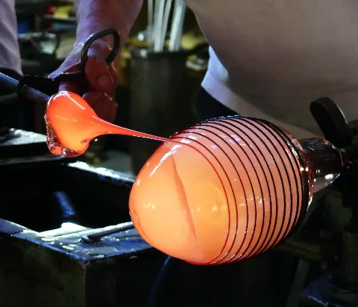 Ein Glasbläser formt gerade eine glühende, orangefarbene Glasmasse zu einem kunstvollen Objekt mit Streifenmuster.