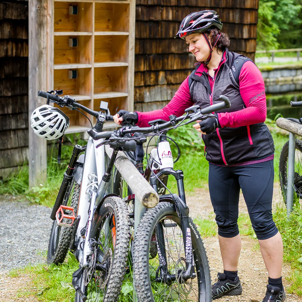Eine Radfahrerin in sportlicher Kleidung überprüft ihr Mountainbike, das Sie an einem Holzständer vor der Racheldiensthütte abgestellt hat.
