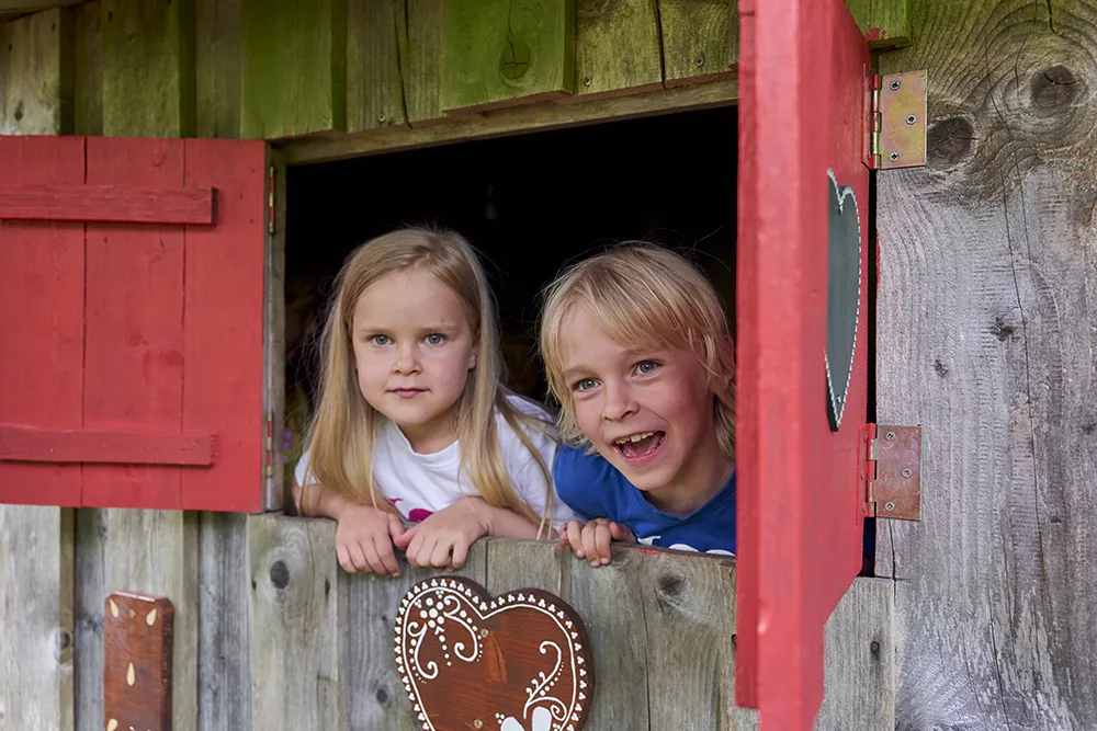 Zwei Kinder schauen fröhlich aus einem Holzfenster mit roten Läden, aus einem Haus mit Pfefferkuchendeko.