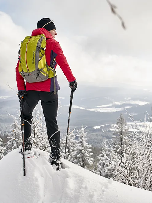 Ein Wanderer in roter Jacke und grünem Rucksack steht auf einem schneebedeckten Felsen und blickt auf die winterliche Landschaft der Ferienregion Nationalpark Bayerischer Wald