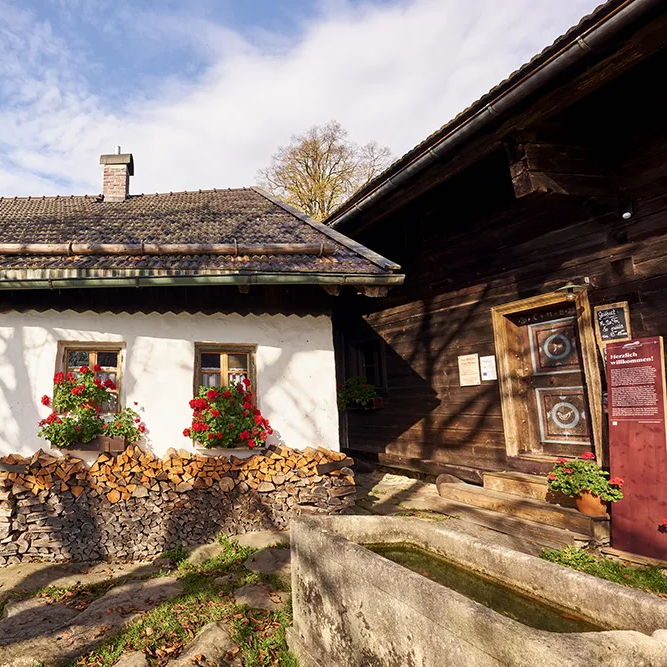 Zwei historisch typische Bayerwald-Häuser stehen dicht beieinander, im Bauernhausmuseum, umgeben von bunten Blumen und Holzstapeln davor im Hofbereich.